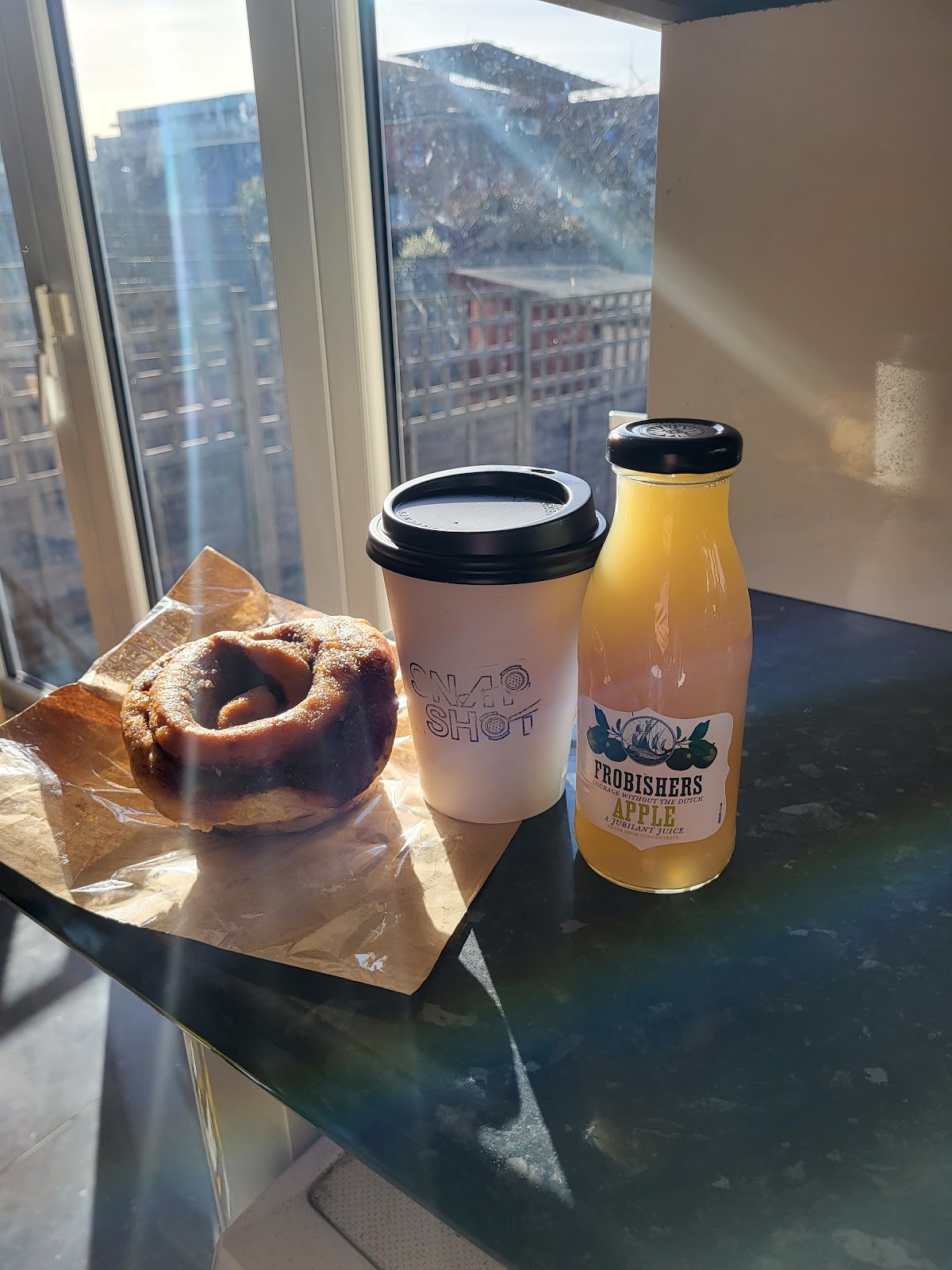 A cinnamon pastry on paper beside a takeaway cup and a bottled orange juice